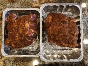 Two rubbed pork butts on wire racks in disposable foil pans