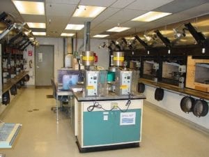 The division lab burn room. The two machines on the table in the foreground apply a measured amount of solvent to each charcoal sample prior to lighting. Charcoal rings are seen hanging below each test station. Not shown in this photo is a special test station that burns charcoal on a scale so weight can be measured throughout the burn test.